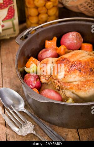 Gebratenes Huhn im holländischen Stil mit gebackenen roten Kartoffeln, Karotten und Sellerie, in einem gusseisernen Topf auf einem rustikalen Holztisch, mit einem alten Kochbuch und einer Konserve Stockfoto