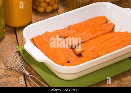 Gebackene Süßkartoffeln in einer rechteckigen Backform auf einer rustikalen Holz-Tischplatte mit antiker Schneebesen und Konserven im Hintergrund Stockfoto