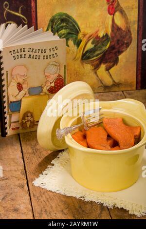 Gebackene kandierte Süßkartoffeln in einer antiken runden Kasserolenschale, die auf einer StoffServiette ruht, auf einer rustikalen Holztischplatte mit einem alten Kochbuch und Huhn Stockfoto