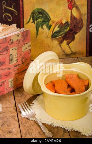 Gebackene kandierte Süßkartoffeln in einer antiken runden Kasserolenschale, die auf einer StoffServiette ruht, auf einer rustikalen Holztischplatte mit einem alten Kochbuch und Huhn Stockfoto