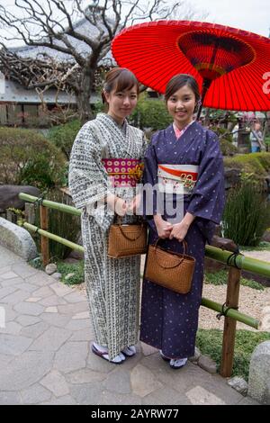 Zwei junge japanische Frauen, die traditionelle Kimonos tragen, posieren vor einem roten Sonnenschirm im Hase Kannon-Tempel (buddhistischer Tempel) in Kamakura, Kanagaw Stockfoto