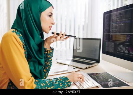 Schöne junge Programmiererin in Hijab, die Code auf dem Computerbildschirm überprüft und Sprachnachrichten für Kollegen aufzeichnet Stockfoto