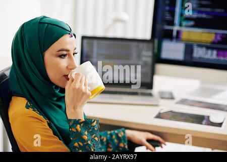 Schöne junge muslimin in Hijab, die am Bürotisch sitzt und einen Becher Kaffee trinkt Stockfoto