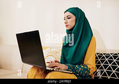 Muslimische, freiberufliche Programmiererin, die zu Hause auf dem Sofa sitzt und am Laptop arbeitet Stockfoto