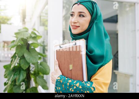 Portrait junger, hübscher muslimischer Geschäftsfrau im Freien mit Zwischenablage und Ordner Stockfoto