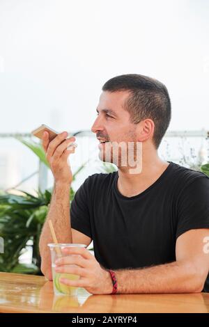 Gutaussehender junger Mann, der erfrischendes Zitrugetränk trinkt und Sprachnachrichten aufzeichnet Stockfoto