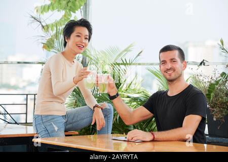 Fröhlicher junger Mann und Frau, die mit einer Brille erfrischender Zitrucocktails toasten und die Kamera betrachten Stockfoto