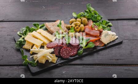 Food-Bannerformat. Verschiedene Käsesorten, Würstchen, geräuchertes Fleisch, Salat, Brot und Oliven auf einer dunklen flachen Tuffplatte auf einem dunklen Holztisch. Meterran Stockfoto