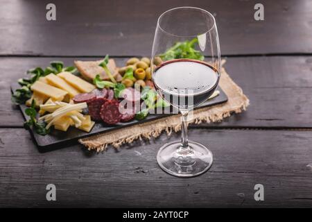 Verschiedene Käsesorten, Würstchen, geräuchertes Fleisch, Salat, Brot, Oliven, Glas Rotwein und auf einem dunklen flachen Teller Tuff auf einem dunklen Holztisch. Meterran Stockfoto