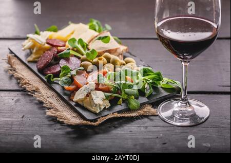 Verschiedene Käsesorten, Würstchen, geräuchertes Fleisch, Salat, Brot, Oliven, Glas Rotwein und auf einem dunklen flachen Teller Tuff auf einem dunklen Holztisch. Meterran Stockfoto