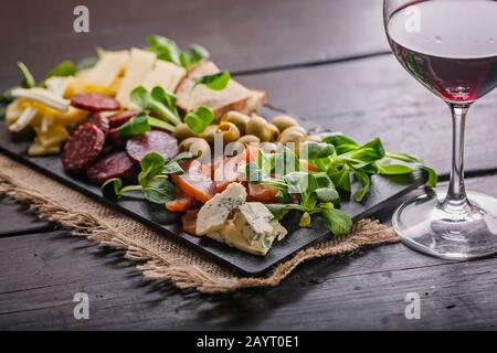 Köstliche und leckere mediterrane Snacks. Verschiedene Käsesorten, Würstchen, geräuchertes Fleisch, Salat, Brot, Oliven, Glas Rotwein und auf einem dunklen flachen Teller Stockfoto