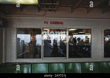 Reflexionen, Pendler und draußen grüne Kunststoffsitze, die durch die Fenster der Staten Island Ferry blicken, lackierter Stahl, ein Rauchverbot Stockfoto