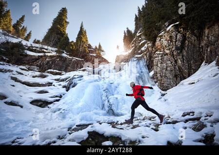 Alter Läufer mit grauem Bart und roter Jacke, der im Winter in der Nähe eines gefrorenen Wasserfalls in den Bergen springt. Sky Running und Trail Running Outdoor-Aktivitäten Co Stockfoto