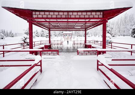 Frau in weißer Jacke spaziert im japanischen Wintergarten mit roter Pagode bei starkem Schneefall im Stadtpark von Almaty, Kasachstan Stockfoto