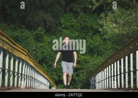 Aktiver junger bärtiger Mann im schwarzen Trikot und graue Shorts, die auf einer Holzbrücke im grünen Park spazieren gehen, im Freien, Freizeitsport Stockfoto