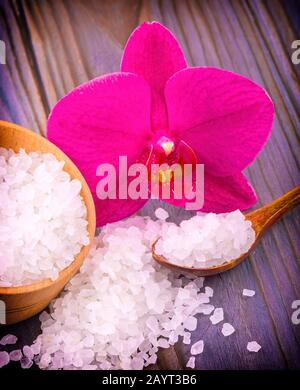 Das weiße Bad Salz in eine hölzerne Schale mit einem Löffel auf einen hölzernen Tisch mit einer Orchidee Stockfoto