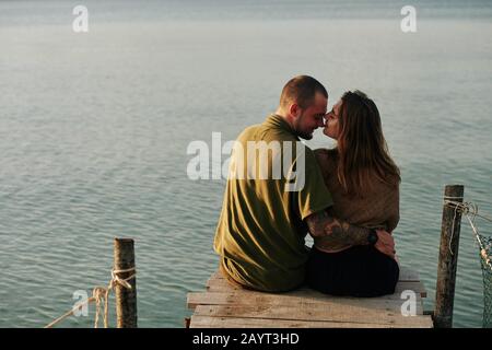 Das küssende junge Paar sitzt gern auf dem Holzsteg und hat einen Blick von hinten Stockfoto