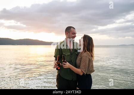 Fröhliches junges Paar hält Bierflaschen und schaut sich beim stehen am Strand mit Liebe an Stockfoto