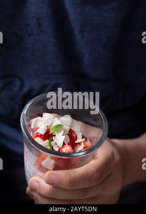 Mann hält mit der Hand ein Glas Erdbeerstücke, gemischt mit Kokospänen, Honig, chia und Milch. Stockfoto