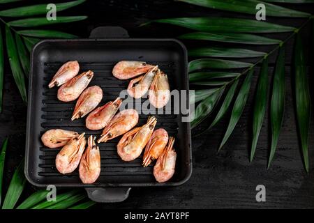 Gegrillte Garnelen in einer Pfanne mit Rosmarinspring zum Braten. Auf dem Substrat befinden sich grüne Palmblätter. Auf schwarzem Holzhintergrund. Kopierbereich Stockfoto