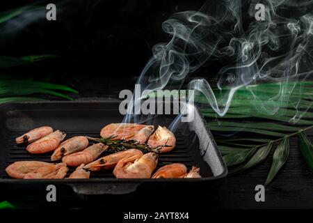 Gegrillte Garnelen in einer Pfanne mit Rosmarinspring zum Braten. Auf dem Substrat befinden sich grüne Palmblätter. Auf schwarzem Holzhintergrund. Kopierbereich Stockfoto