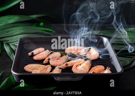 Gegrillte Garnelen in einer Pfanne mit Rosmarinspring zum Braten. Auf dem Substrat befinden sich grüne Palmblätter. Auf schwarzem Holzhintergrund. Kopierbereich Stockfoto