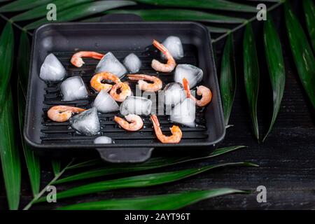 Gegrillte Garnelen in einer Pfanne mit Rosmarinspring zum Braten. Auf dem Substrat befinden sich grüne Palmblätter. Auf schwarzem Holzhintergrund. Kopierbereich Stockfoto