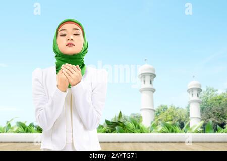Asiatische Muslimin in einem Schleier stehend, während sie die Hände hob und auf der Moschee-Terrasse betete Stockfoto