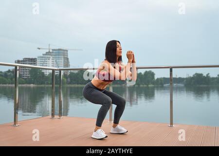 Schöne Dame mit muskulösem Körper in Sportbekleidung, die auf einem Holzsteg in der Nähe des Sees hocken wird. Athletin mit dunklem Haartraining an frischer Luft. Stockfoto