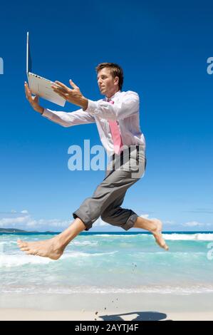 Energiegeladener Büroangestellter springt mit seinem Laptop an einem leeren tropischen Strand entlang Stockfoto