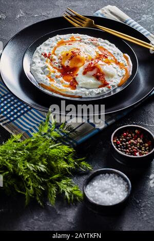 Traditionelles türkisches Frühstück, Cilbir, pochierte Eier in Joghurt, gefüllt mit würziger Butter, die auf schwarzen Tellern mit goldenem Besteck serviert wird, Verti Stockfoto