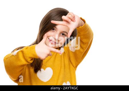 Entzückendes Mädchen im Elementaralter, das lächelt, während er die Kamera betrachtet und die Handgeste des Bilderrahmens macht. Studio auf weißem Hintergrund aufgenommen. Stockfoto