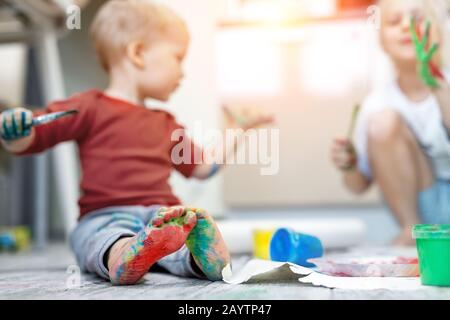 Hübsche süße kaukasische kleine blonde Geschwister Kinder genießen Spaß beim Malen mit Pinsel und Handfläche zuhause. Fröhliche glückliche Kinder lächeln Stockfoto