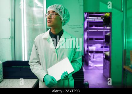 Taille oben Porträt junger asiatischer Arbeiter, die Schutzkleidung tragen, die im Biolabor steht, das von grünem Licht beleuchtet wird, Kopierraum Stockfoto