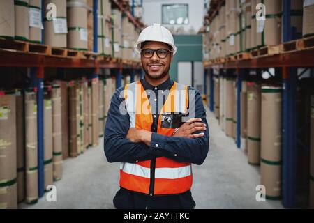 Portrait lächelnder Arbeiter mit orangefarbener Weste und weißem Helm mit Armen, die in einem großen Lagerhaus gekreuzt wurden, lächelnd Stockfoto