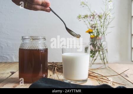 Die Hand der Frau, die etwas natürlichen Honig auf ein Glas Milch auf einen Holztisch in einer rustikalen Küche gegen Blumenschmuck gießt. Leeren Sie den Kopierbereich. Stockfoto