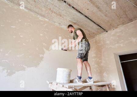 Der Bauherr steht auf einem Holzständer und arbeitet in kleinem Raum mit einem Spachtel mit Putz an der Wand. Wände in Innenräumen putzen. Ein Kerl mit Bart in einem T-Shirt und einer Jeans ist verschmierte Farbe Stockfoto