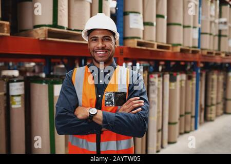 Selbstbewusster afroamerikanischer Mann, der mit gefalteten Armen in der Fertigungseinheit mit weißem Helm und Uniform im Werk steht Stockfoto