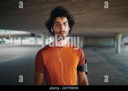 Porträt eines ernsthaften jungen Mannes mit Ohrhörern im Ohr, der unter der Brücke steht und die Kamera betrachtet Stockfoto