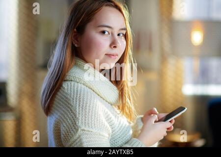 Modernes Teenager-Mädchen mit roten Haaren in weißem Pullover mit Smartphone in sozialen Medien im modernen Haus im sonnigen Wintertag. Stockfoto