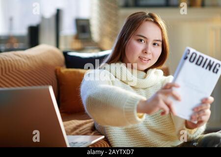 Fröhliches modernes Teenager-Mädchen mit roten Haaren in weißem Pullover mit Laptop, das im modernen Haus am sonnigen Wintertag ein pädagogisches Codierbuch zeigt. Stockfoto