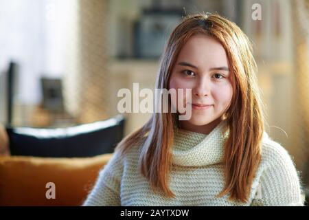 Porträt eines fröhlichen modernen Teenager-Mädchens mit roten Haaren im weißen Pullover im modernen Zuhause am sonnigen Wintertag. Stockfoto