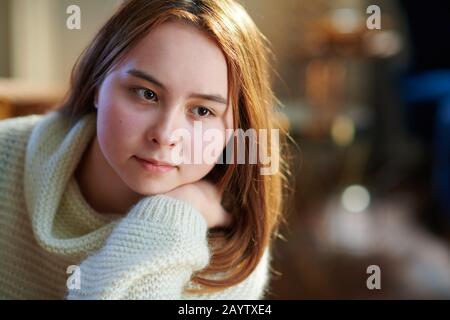 Porträt der nachdenklichen modernen jungen Frau mit roten Haaren im weißen Pullover im modernen Zuhause am sonnigen Wintertag. Stockfoto