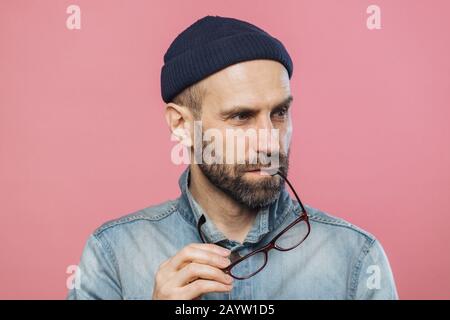 Portrait von nachdenklich, bärtiger Mann zieht Gläser, trägt Denim stilvolle Jacke und Hut, über rosa Hintergrund isoliert. Im mittleren Alter unrasierten Mann contem Stockfoto