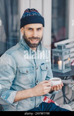 Vertikaler Schuss von attraktivem blauäugigen bärtigen Männchen in stilvoller Kleidung, trinkt heißen Tee, wirkt mit zufriedener Ausdrucksweise bei der Kamera, verbringt Freizeit tim Stockfoto