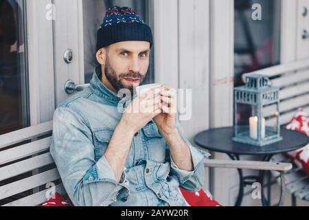 Schuss aus attraktivem Männer mittleren Alters mit dickem dunklen Bart und Schnurrbart, mit blauen Augen, Heißgetränk, gekleidet mit jean-jacke, wirkt direkt drinnen Stockfoto