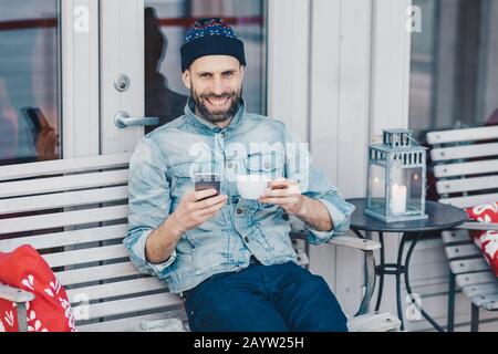 Portrait von bärtigen Männern mit breitem Lächeln, sitzt auf der Bank, verwendet modernes Handy, surfes soziales Netzwerk, freut sich, gute Nachrichten zu erhalten, Getränke aromatisch Stockfoto