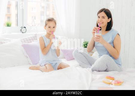 Fröhliche Mutter und Tochter gekleidet im Schlafanzug, Frühstück am Morgen, trinken Milch Shake mit Donuts, gekreuzten Beinen sitzen auf komfortablen Bett in Wh Stockfoto