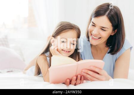 Hübsche Frauen Mutter und ihre kleine Tochter lesen Sie interessantes Buch im Schlafzimmer, auf comfortabled Bett liegen während der Morgen. Schöne Mama liest Märchen für fairty Stockfoto