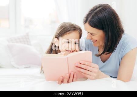 Innen- schuss von angenehmen Suche weibliches Kind mit neugierigen Ausdruck, liest interessante Buch zusammen mit Ihrer liebevollen Mutter, Blick bunte Pi Stockfoto